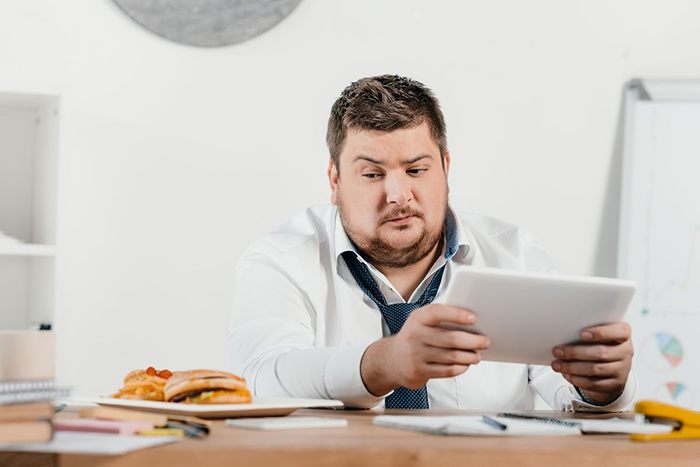 overweight businessman looking at junk food while working with tablet at workplace